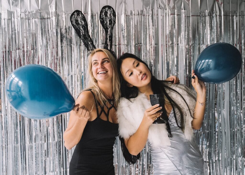 Two women in festive outfits holding balloons at a party with foil curtains.
