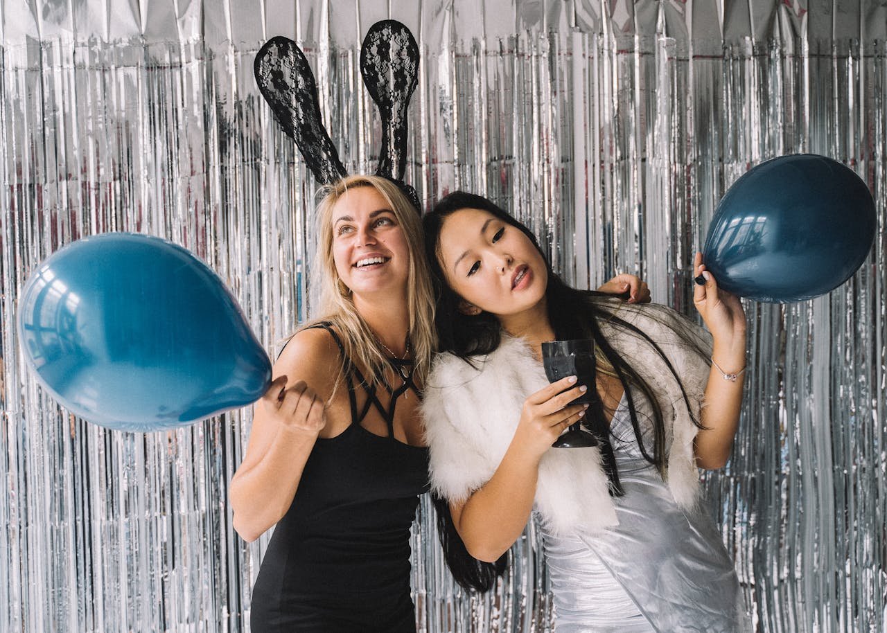 Two women in festive outfits holding balloons at a party with foil curtains.